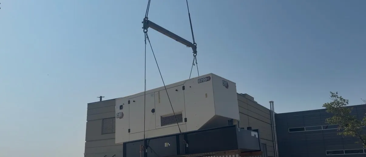 A CAT D200 generator set suspended from a crane boom, being lowered onto a rooftop mechanical deck.